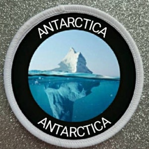 Può includere: Un cerotto rotondo bianco con testo nero e un cerchio nero al centro. Il testo dice "ANTARCTICA" nella parte superiore e inferiore del cerchio. Il cerchio contiene una foto di un iceberg parzialmente sommerso in acqua blu.