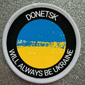Può includere: Un cerotto circolare bianco con testo nero che recita "Donetsk sarà sempre Ucraina" e una bandiera ucraina blu e gialla al centro.