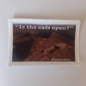 Może przedstawiać: Prostokątna naszywka z białym obramowaniem przedstawia krajobraz i tekst "Is the cafe open?" oraz "Snowdon". Obraz przedstawia brązowy łańcuch górski i budynek.