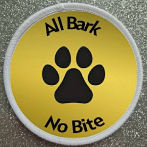 Puede incluir: Un parche redondo blanco con fondo amarillo y texto negro y una huella de pata negra. El texto dice "All Bark No Bite".