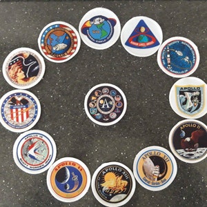 May include: A collection of 12 round patches featuring the Apollo missions. Each patch has a different design with a white background and colorful details. The patches are arranged in a circle on a dark gray surface.