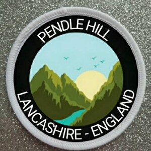Può includere: Una toppa rotonda con la scritta "PENDLE HILL LANCASHIRE - ENGLAND". Il disegno raffigura un paesaggio con montagne verdi, un fiume blu, un sole giallo e un cielo blu con uccelli. Ha un bordo nero e un bordo cucito bianco.