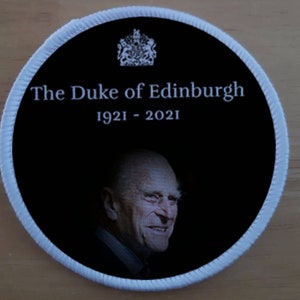 May include: A black and white round patch with a white border. The patch features a portrait of Prince Philip, Duke of Edinburgh, with the text "The Duke of Edinburgh 1921 - 2021" above the portrait.