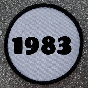 Op de afbeelding: Ronde witte patch met zwarte stiksels en het jaar 1983 in het zwart gedrukt.
