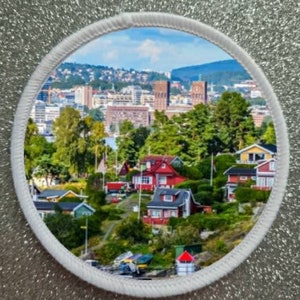 May include: A circular patch with a white border and a colorful image of a city skyline with red-roofed houses in the foreground. The city skyline features tall buildings with a blue sky and white clouds in the background.