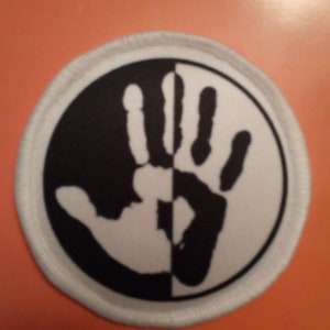 Könnte beinhalten: Ein runder weißer und schwarzer Patch mit einem Bild von zwei Händen, eine schwarz und eine weiß, die sich gegenüberstehen. Die Hände sind Spiegelbilder voneinander.