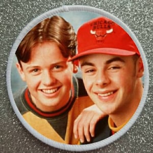 May include: A white round patch with a photo of two boys. The boy on the right is wearing a red Chicago Bulls baseball cap. The boy on the left is wearing a yellow and green shirt. The text "Chicago Bulls" is on the cap.