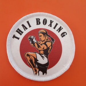 Puede incluir: Un parche redondo blanco con un círculo rojo en el centro. El círculo tiene una ilustración en negro y naranja de un luchador de Muay Thai. El texto "THAI BOXING" está escrito en negro alrededor del círculo rojo.