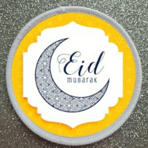 May include: A white round sticker with a yellow border and a crescent moon design. The moon has a blue and white geometric pattern. The text "Eid Mubarak" is written in black cursive lettering.