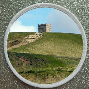 Pode incluir: Um remendo circular branco com uma foto de um edifício de pedra em uma colina gramada. O edifício tem um telhado pontudo e está localizado em uma colina com um caminho que leva até ele.