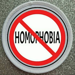 Op de afbeelding: Een ronde patch met een witte kern en het woord "HOMOPHOBIA" in zwart, doorgestreept door een rode diagonale lijn. De patch heeft een witte rand met een zwarte omtrek en een grijs gestikte rand.
