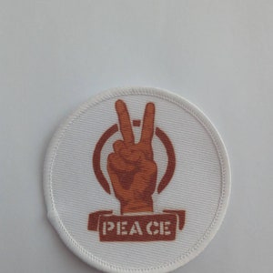 Puede incluir: Un parche redondo blanco con un símbolo de paz marrón y la palabra "PEACE" escrita debajo.