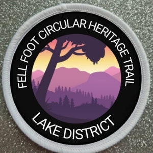 May include: A black and white circular patch with the text "Fell Foot Circular Heritage Trail Lake District" around the edge. The centre of the patch features a silhouette of a mountain range with a tree in the foreground.