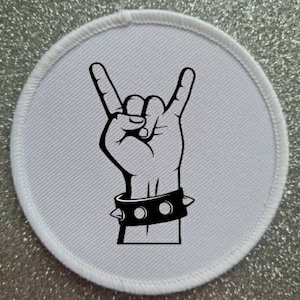 May include: A white, circular patch with a black illustration of a hand making the rock and roll gesture. The hand is wearing a black studded wristband. The patch has a white stitched border.