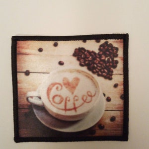 May include: A brown and black embroidered patch featuring a white coffee cup with a heart and the word "coffee" written in brown on the foam. The cup is on a wooden surface with coffee beans scattered around it.