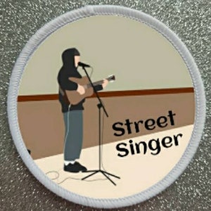 May include: A white round patch with a brown background and a black outline. The patch features a cartoon illustration of a person playing a guitar with the text "Street Singer" in black.