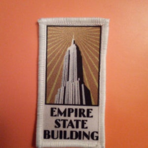 Op de afbeelding: Een rechthoekige patch met het Empire State Building. Het gebouw is afgebeeld in wit en zwart tegen een goud-bruine achtergrond met stralende lijnen. De woorden "EMPIRE STATE BUILDING" staan in het zwart onderaan gedrukt, omlijst door een witte rand.