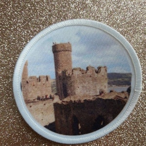 Könnte beinhalten: Ein weißer runder Aufnäher mit einem aufgedruckten Bild einer braunen Steinburg. Die Burg hat einen hohen Turm und ist von einer Mauer umgeben. Der Himmel ist blau mit weißen Wolken.