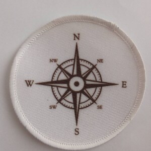 May include: White fabric patch with a brown compass design. The compass has the cardinal directions N, S, E, and W, as well as the intermediate directions NE, NW, SE, and SW.