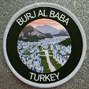 Può includere: Un cerotto rotondo bianco e nero con il testo "Burj Al Baba Turchia" in lettere bianche. Il cerotto presenta una foto di una città con edifici bianchi e un paesaggio verde.