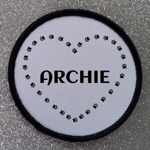 May include: White round patch with a black border. The patch has a black paw print heart design with the name "ARCHIE" in the center.