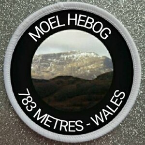 May include: A white and black round patch with the text "Moel Hebog 783 Metres - Wales" and a photo of a mountain range in the center.