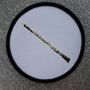 May include: A white embroidered patch with a black border, featuring a black and silver oboe.