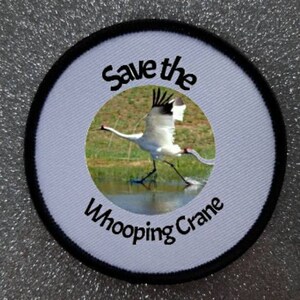 May include: White embroidered patch with black text that reads "Save the Whooping Crane". The patch features a photo of a white whooping crane in flight over a grassy area with water.