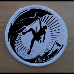 Puede incluir: Un parche redondo blanco con una silueta negra de un escalador ascendiendo una pared de roca. El escalador está en silueta sobre un fondo blanco con rayos negros que emanan del centro. El parche tiene un borde blanco con costuras negras.