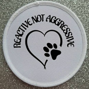May include: White round patch with black text that reads "Reactive Not Aggressive" with a black paw print inside a heart.