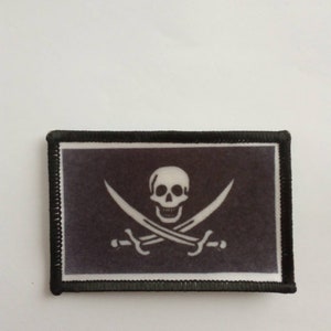 May include: A black and white embroidered patch with a skull and crossbones design. The skull is white with black eyes and a black mouth. The crossbones are white and are crossed behind the skull. The patch is surrounded by a black border.