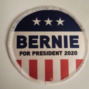 Può includere: Un bottone circolare bianco, blu e rosso con il testo "Bernie for President 2020" e tre stelle bianche.