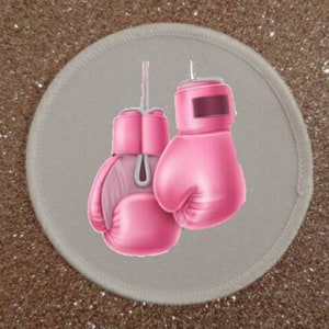 May include: A pair of pink boxing gloves hanging on a white circular patch. The gloves are tied together with a white string.