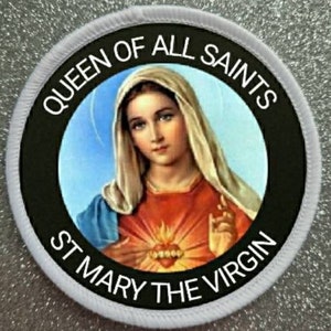 May include: White circular patch with black text and a black circle in the center. The text reads "Queen of All Saints St Mary the Virgin". Inside the black circle is a color image of the Virgin Mary with a blue robe and a red and gold heart.