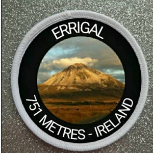 May include: White round patch with black border and white text. The text reads "Errigal 751 Metres - Ireland". A photo of a mountain is inside the circle.