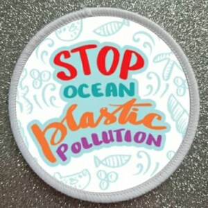 Op de afbeelding: Ronde witte patch met een blauwe achtergrond en kleurrijke tekst die "STOP OCEAN PLASTIC POLLUTION" zegt. De achtergrond is versierd met een witte omtrek van vissen, golven en andere afbeeldingen die met de oceaan te maken hebben.