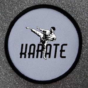 Puede incluir: Un parche redondo blanco con un borde negro. El parche presenta una silueta en blanco y negro de una persona realizando una patada de karate. El texto "KARATE" está impreso en negro debajo de la silueta.
