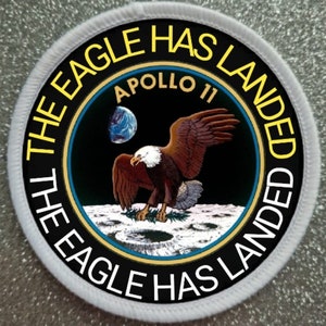 Pode incluir: Um remendo circular branco com texto preto e amarelo que diz "The Eagle Has Landed The Eagle Has Landed" ao redor da borda. No centro há um círculo preto com o texto "Apollo 11" e um gráfico de uma águia pousando na lua com a Terra ao fundo.