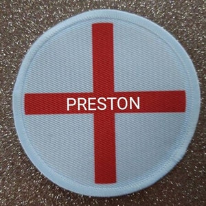 Puede incluir: Un parche redondo blanco con una cruz roja y la palabra "PRESTON" en el centro.