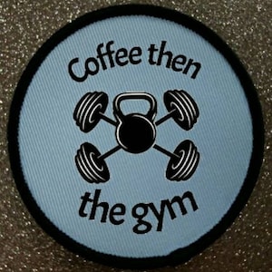 Puede incluir: Un parche redondo azul claro con texto negro y un borde negro. El texto dice "Coffee then the gym". Una pesa rusa negra y cuatro pesas negras están en el centro.