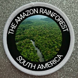 Könnte beinhalten: Ein weißer runder Patch mit schwarzem Rand und weißem Text, der "The Amazon Rainforest South America" lautet. Die Mitte des Patches zeigt ein Foto eines üppigen, grünen Regenwaldes mit einem Fluss, der sich durch ihn schlängelt.
