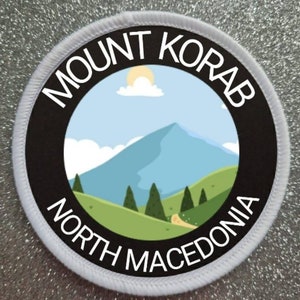 May include: White round patch with black border and text "MOUNT KORAB NORTH MACEDONIA". The center features a cartoon illustration of a mountain range with a blue sky and white clouds.