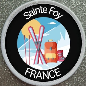 May include: A black and white circular patch with the text "Sainte Foy FRANCE" in white. The center of the patch features a colorful illustration of a mountain scene with skis, ski poles, a backpack, and ski boots.