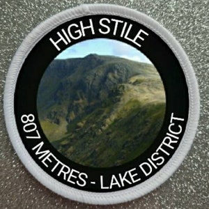 May include: White round patch with black text and a photo of a mountain range. The text reads "High Stile 807 Metres - Lake District".