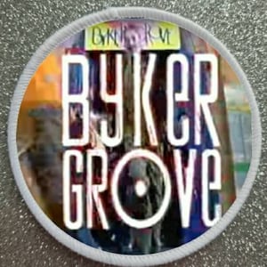 May include: Round patch with the words "Byker Grove" in large white capital letters. The patch has a white border and a colorful background. The text "Byker Grove" is in a bold, sans-serif font.
