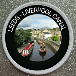 May include: A white and black round patch with the text "Leeds-Liverpool Canal" in white lettering. The patch features a photo of a canal with boats and buildings.