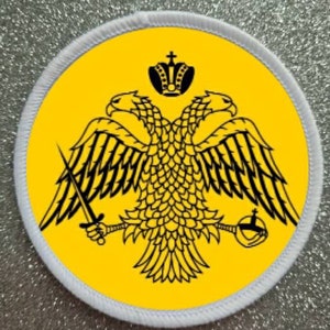 May include: A white circular patch with a yellow background featuring a black double-headed eagle with a crown and a sword. The eagle is a symbol of the Byzantine Empire.