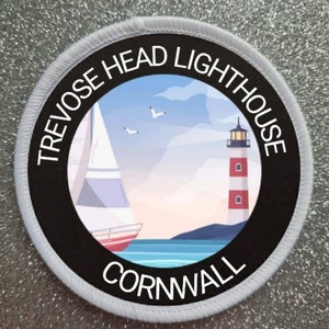 May include: A white and black round patch with the text "Trevose Head Lighthouse Cornwall" around the edge. The center of the patch features a colorful illustration of a lighthouse, a sailboat, and seagulls against a blue sky.
