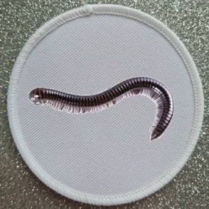 May include: A round, white patch with a stitched border, featuring a detailed illustration of a millipede. The millipede is dark brown and black, curved across the patch. The background is a light gray fabric.