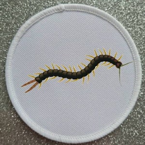 May include: Embroidered patch featuring a centipede design. The centipede is depicted in shades of brown and yellow, with a detailed, realistic appearance. The patch is circular, with a white background and a white stitched border.